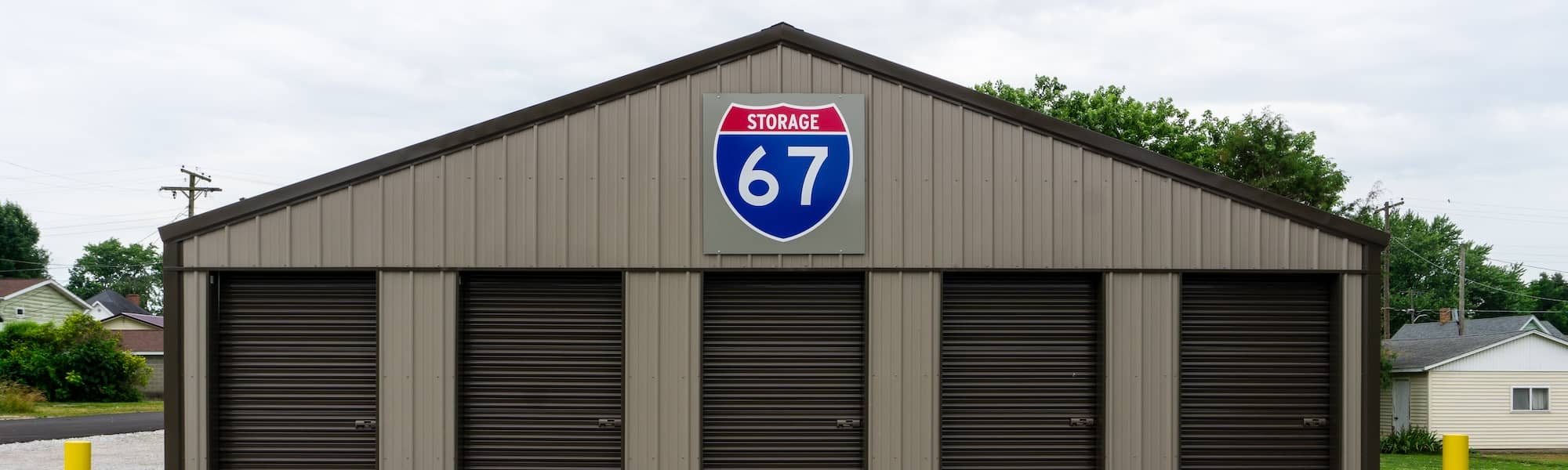 Front view of 67 Storage facility in Worthington with brown metal exterior and five roll-up doors, featuring a prominent blue and red highway shield sign with the number 67.